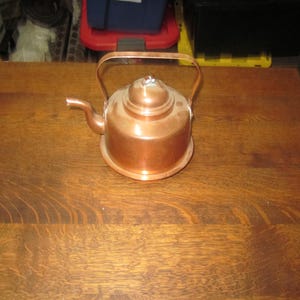 May include: A copper tea kettle with a handle and spout. The kettle has a round base and a lid with a decorative knob. The copper has a shiny, reflective surface. The kettle is sitting on a wooden surface.