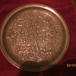 May include: A round, copper-coloured metal tray with a raised design of a scene with people and animals. The tray has a smooth, shiny surface.