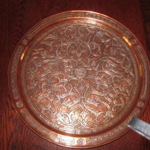 Könnte beinhalten: Ein rundes Serviertablett aus Kupfer mit einem aufwendigen silbernen Design aus stilisierten floralen Mustern. Das Tablett hat einen erhöhten Rand mit einem dekorativen Rand.