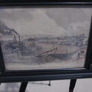 May include: A framed black and white print of a river scene with a steamboat, buildings, and figures. The print is in a black frame and is displayed on a black easel.