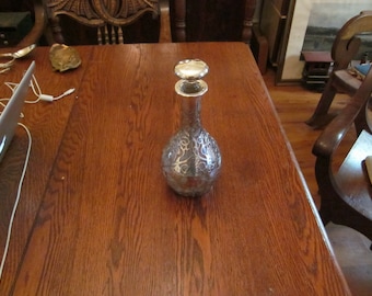May include: A silver decanter with a clear glass body and a silver stopper. The decanter has an intricate design etched into the glass. The decanter is sitting on a wooden table.