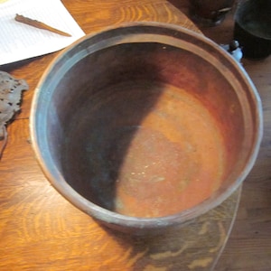 May include: A large copper pot with a worn, aged patina. The inside of the pot is stained with a light brown and blue discoloration.