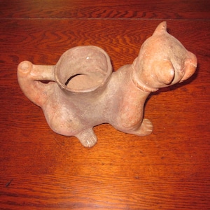 Pode incluir: Uma escultura de cerâmica de terracota de um gato com uma grande boca aberta. O gato está sentado em uma superfície de madeira e tem um espaço redondo e oco nas costas que pode ser usado como um vaso ou um vaso.