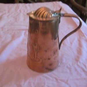 May include: A copper pitcher with a hammered finish and a decorative lid. The pitcher has a handle and a floral design.