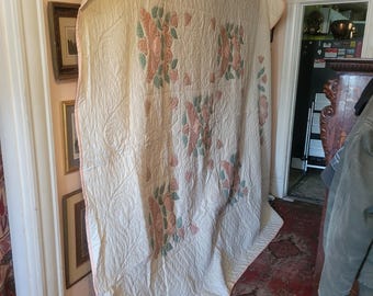 May include: A white quilt with a pink and green floral pattern. The quilt is hanging on a wooden rod and is folded in half. The quilt is in a room with a pink wall and a wooden floor.