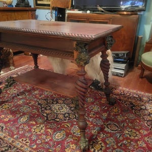 May include: An antique dark wood table with a rectangular top and a lower shelf. The table features ornate carvings on the legs and corners, with a twisted leg design. It stands on a patterned red rug.
