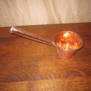 May include: A copper ladle with a long handle and a rounded bowl. The ladle is made of shiny copper and sits on a wooden surface. The handle extends from the bowl, creating a functional and decorative kitchen tool.