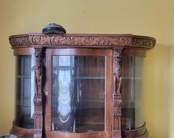 May include: An antique wooden display cabinet with glass doors and ornate carvings. The cabinet has a curved front and sits on a wooden base with drawers. A hat rests on top. Various objects are on the cabinet's surface.