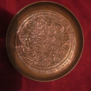 May include: A round, copper-coloured tray with an intricate embossed design of flowers, birds, and animals. The tray has a raised rim and a smooth, polished surface.