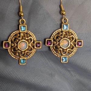 May include: A pair of gold-toned earrings with a circular design. Each earring features a central opal-like stone, surrounded by a floral pattern, and accented with square blue and purple gemstones. The earrings have a hook closure.