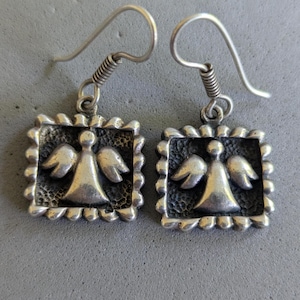 May include: A pair of silver-toned drop earrings featuring a square frame with a scalloped edge. Each earring displays a raised angel figure centered against a dark, textured background. The earrings are attached to simple silver-toned ear wires.