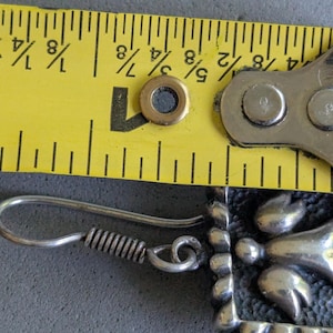 May include: A silver-toned earring featuring a square frame with a textured dark background and a central angel figure. A yellow measuring tape is positioned above the earring, indicating a width of approximately 0.75 inches for the square component.