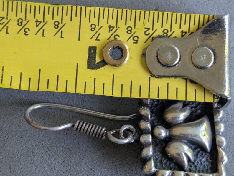 May include: A silver-toned earring featuring a square frame with a textured dark background and a central angel figure. A yellow measuring tape is positioned above the earring, indicating a width of approximately 0.75 inches for the square component.