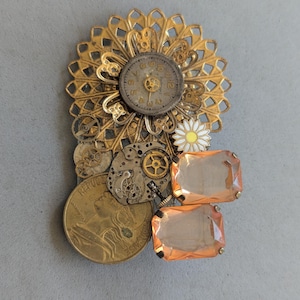 May include: A brooch featuring a gold-toned filigree sunburst, a circular watch face with numerals, a small white and yellow daisy charm, various metallic clockwork gears, a gold-colored coin with a profile portrait, and two rectangular peach crystals.