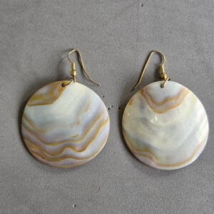 May include: A pair of circular drop earrings featuring polished mother-of-pearl discs with natural, swirling bands of iridescent white, cream, and golden-brown hues. Each disc is suspended from a simple gold-toned metal hook wire for attachment.