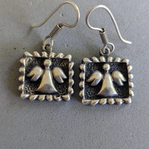 May include: A pair of silver-toned drop earrings featuring a square frame with a scalloped edge. Each earring displays a raised, central angel figure with wings against a dark, textured background. The earrings hang from simple silver-toned hooks.