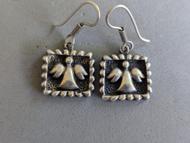 May include: A pair of silver-toned drop earrings featuring a square frame with a scalloped edge. Each earring displays a raised, central angel figure with wings against a dark, textured background. The earrings hang from simple silver-toned hooks.