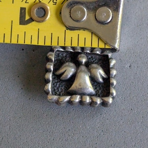 May include: A square silver-toned metal charm featuring a raised angel figure with wings against a textured background, framed by a beaded border. A yellow measuring tape is positioned along the top edge, showing a width of approximately 0.75 inches.