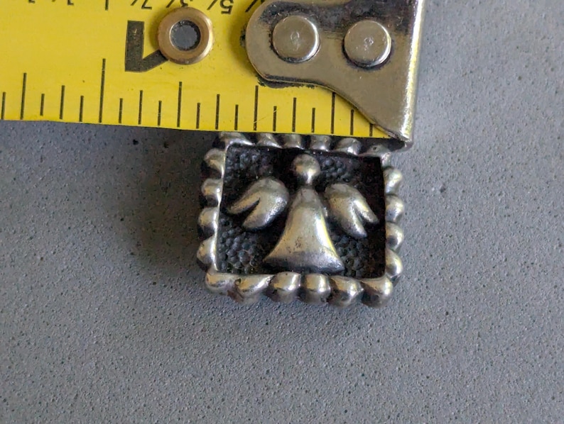 May include: A square silver-toned metal charm featuring a raised angel figure with wings against a textured background, framed by a beaded border. A yellow measuring tape is positioned along the top edge, showing a width of approximately 0.75 inches.