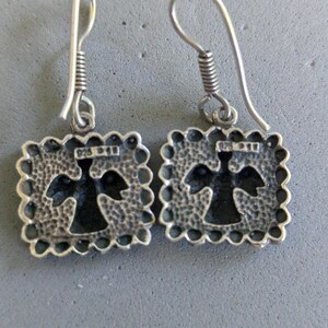 May include: A pair of sterling silver drop earrings featuring square pendants with a scalloped edge and a hammered texture. Each pendant has a central cutout in the shape of an angel and is stamped with 925 on the front. The earrings hang from silver hooks.