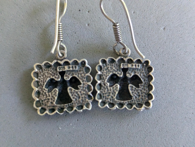 May include: A pair of sterling silver drop earrings featuring square pendants with a scalloped edge and a hammered texture. Each pendant has a central cutout in the shape of an angel and is stamped with 925 on the front. The earrings hang from silver hooks.