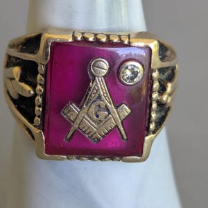 May include: A gold-toned ring with a rectangular, magenta-colored center stone. The stone features a gold Masonic symbol and a small, clear gemstone. The ring has ornate detailing on the band.