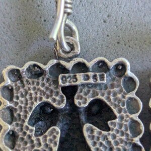 May include: A silver earring featuring a square pendant with a textured, dimpled surface and a scalloped edge. The metal is stamped with the text 925 and SE. A silver hook is attached to the top of the pendant through a small circular loop.