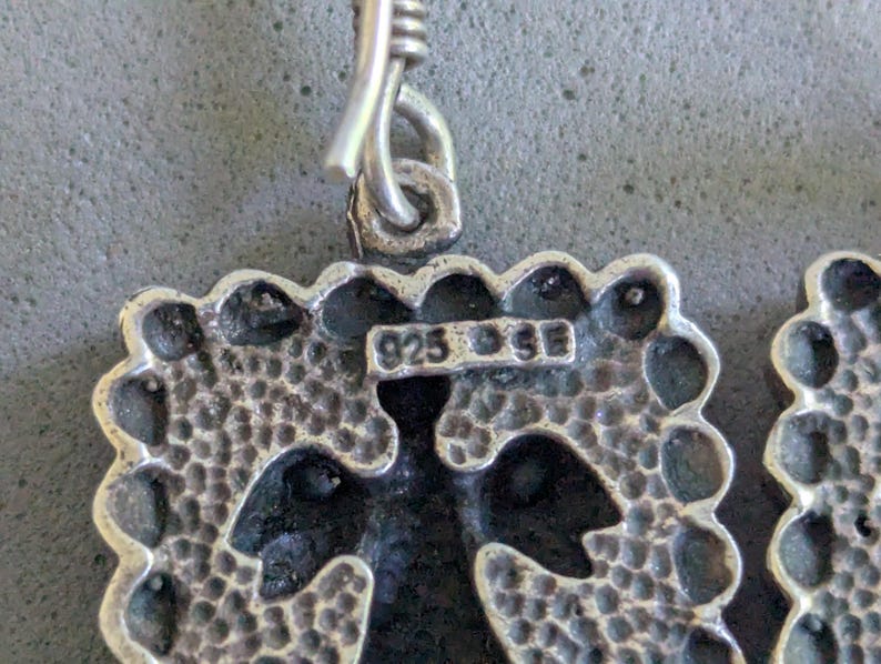 May include: A silver earring featuring a square pendant with a textured, dimpled surface and a scalloped edge. The metal is stamped with the text 925 and SE. A silver hook is attached to the top of the pendant through a small circular loop.