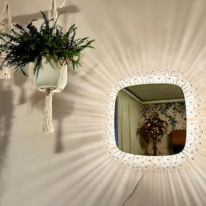 Op de afbeelding: Een witte, vierkante spiegel met een decoratieve, kristal-achtige rand. De spiegel is gemonteerd op een witte muur en reflecteert een kamer met een behang met patronen en een kamerplant.