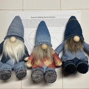 Puede incluir: Tres gnomos de mezclilla sentados en una superficie blanca. Los gnomos llevan sombreros, camisas y pantalones de mezclilla azul. Un gnomo lleva una camisa a cuadros rojos y blancos. Los gnomos tienen barbas blancas y marrones. Los gnomos están hechos a mano y tienen un encanto rústico y campestre.