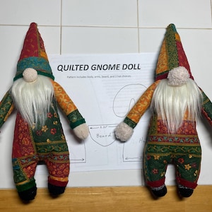 May include: Two quilted gnome dolls with white beards and colorful patchwork fabric. The dolls are wearing red and green hats and have white fabric for hands. The dolls are standing on a white surface with a pattern for making a gnome doll.