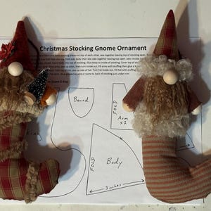 May include: Two Christmas stocking gnome ornaments. One gnome has a red plaid stocking body and a red hat with a flower. The other gnome has a striped stocking body and a plaid hat. Both have a fluffy beard and a wooden nose.