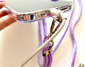 May include: A clear phone case with a lavender phone strap. The strap has a silver clasp and two silver rings. A white charging cable is plugged into the phone.