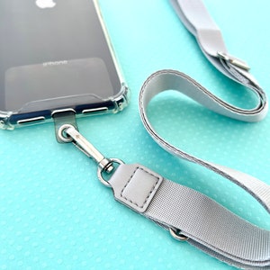 Może przedstawiać: Przezroczyste etui na telefon z przymocowanym szarym paskiem. Pasek ma srebrny klips i srebrny pierścień. Etui na telefon znajduje się na jasnoniebieskim tle.
