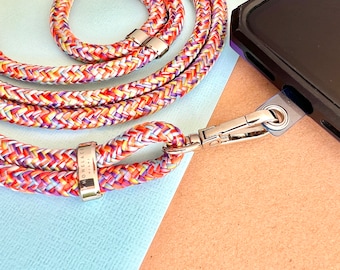 May include: A colorful braided phone strap with a silver clasp and a silver tag that says "Homelace" on it. The strap is on a blue and brown surface.
