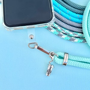 Może przedstawiać: Przezroczyste etui na telefon z miętowym sznurkiem do telefonu. Pasek ma srebrne zapięcie i zawieszkę w kształcie filiżanki kawy. Dodatkowe kolory sznurka to szary, niebieski i turkusowy. Telefon znajduje się na jasnoniebieskim tle.
