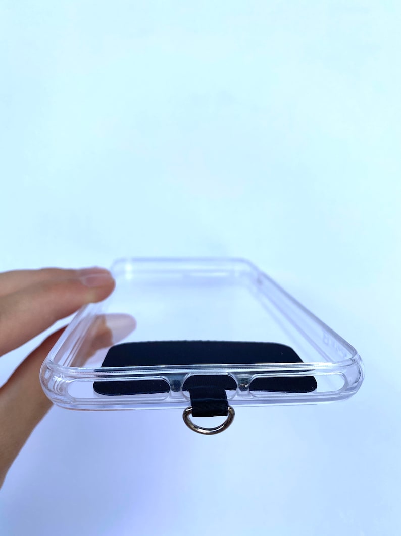 May include: A clear phone case with a black rubber strap and a metal ring. The case has a black rubber insert to protect the phone.