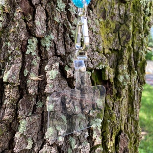 May include: A clear plastic badge holder with a silver clip attached to a light blue lanyard. The lanyard is hanging from a tree trunk.