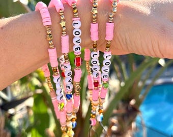 May include: A set of six pink beaded bracelets with gold accents and the word "LOVE" spelled out on each bracelet. The bracelets are layered on a person's wrist.