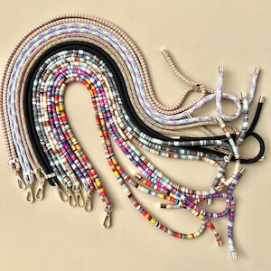 Puede incluir: Una colecci&oacute;n de correas de tel&eacute;fono coloridas con varios patrones y dise&ntilde;os. Las correas est&aacute;n hechas de cuerda y cuentas y tienen cierres de color dorado.
