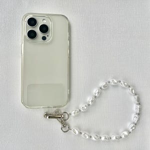 Może przedstawiać: Przezroczyste etui na telefon z perłową bransoletką na nadgarstek. Etui ma przezroczysty tył i wypukłość na aparat. Bransoletka wykonana jest z białych, owalnych pereł i srebrnych metalowych akcentów.
