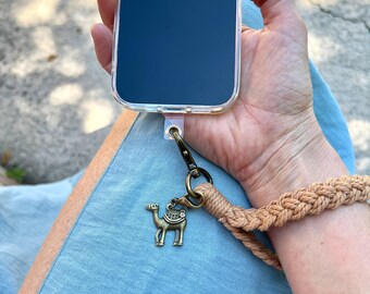 May include: A clear phone case with a camel charm attached to a braided wrist strap. The camel charm is bronze-colored. The wrist strap is a light brown color. The phone screen is dark blue.