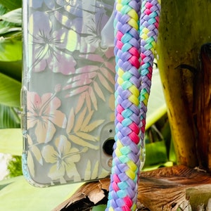 Może przedstawiać: Przezroczyste etui na telefon z kwiatowym wzorem i wielokolorowym plecionym paskiem. Pasek jest wykonany z różowej, żółtej, niebieskiej i zielonej przędzy.