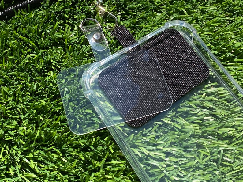May include: A clear phone case with a black adhesive pad on the back. The case is attached to a black lanyard with a silver clasp.