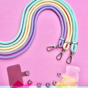 Może przedstawiać: Trzy kolorowe sznurki do zawieszek w pastelowym zielonym, żółtym i fioletowym kolorze, ze srebrnymi klamrami. Sznurki są ułożone w kształt tęczy na różowym tle.