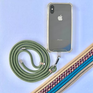 Może przedstawiać: Przezroczyste etui na telefon z iPhonem w środku, przymocowane do zielonej smyczy z metalowymi elementami w kolorze srebrnym. Widoczne są również wielokolorowe, tkane paski, oferujące alternatywne style.