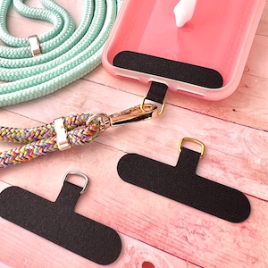 May include: Two black phone strap holders with gold metal rings. A pink phone case with a black phone strap holder attached. A multi-colored braided phone strap with a gold metal clasp.