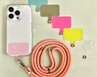 May include: A white iPhone case with a pink accent and a coral phone strap. The case has a clear back and a raised camera bump. The strap is made of a thick, braided cord. The image also shows a variety of colorful stickers.