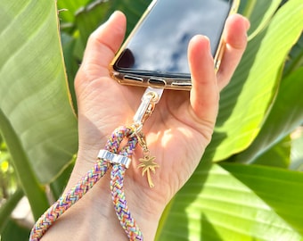 May include: A colorful braided phone strap with a gold palm tree charm attached to a clear phone case. The strap is attached to the phone case with a gold metal ring.