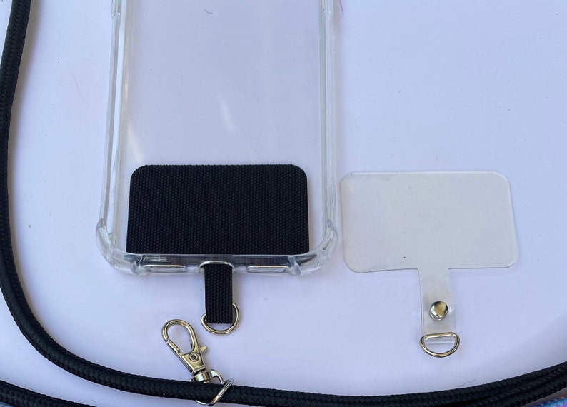 May include: A clear phone case with a black fabric patch on the back. The case has a black lanyard attached to it with a silver clip. A separate clear phone case with a silver clip is also shown.